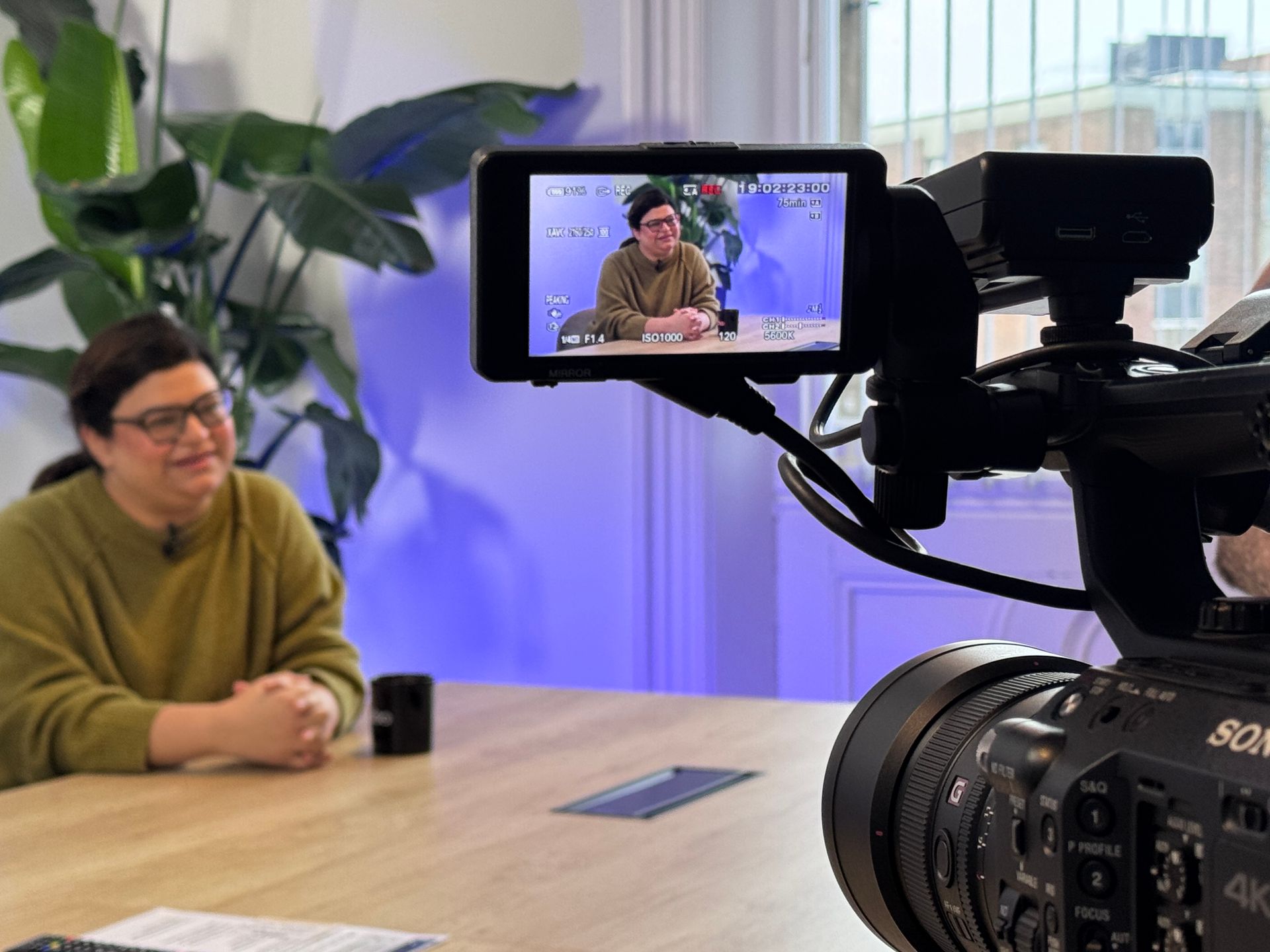 A person in an olive sweater sits at a wooden table in a room with a large plant, viewed through a professional camera.