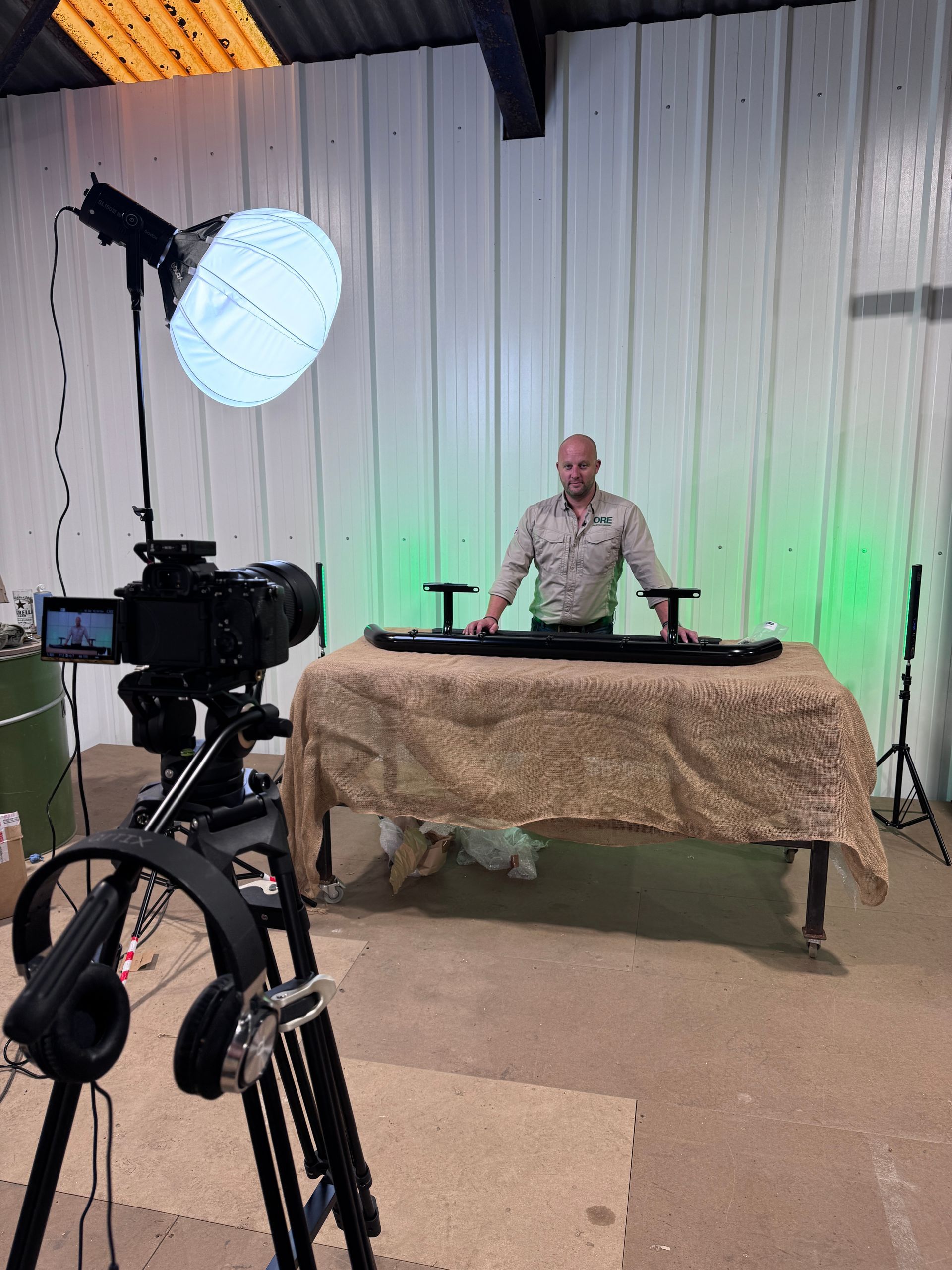 A person standing behind a table with parts in a workshop, filming a video with professional lighting and a tripod camera.