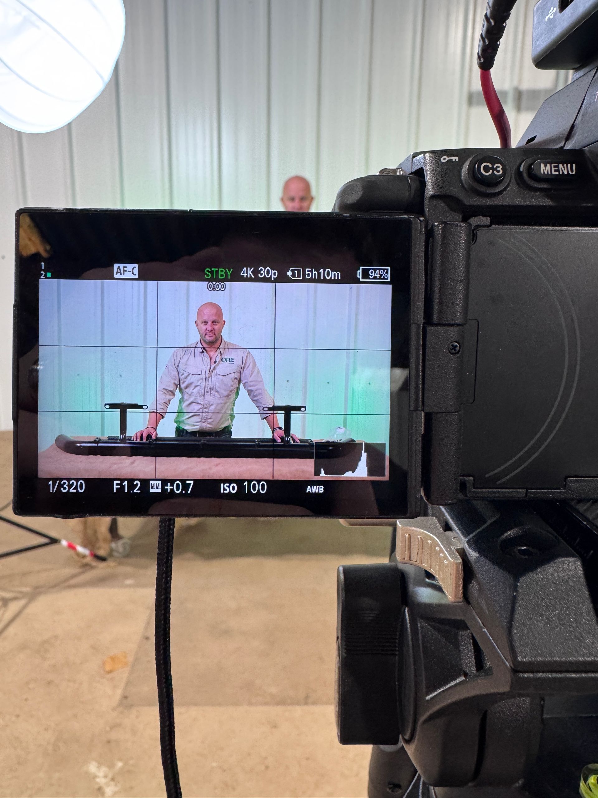 A camera screen shows a person standing behind a desk in a studio, preparing to film a video presentation.