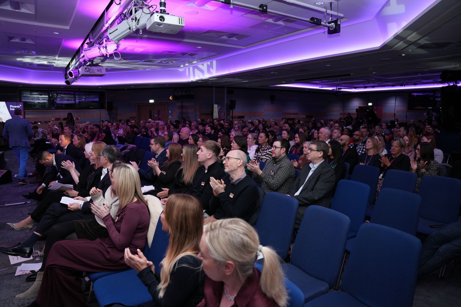 An audience in a conference room with purple lighting, watching a presentation.