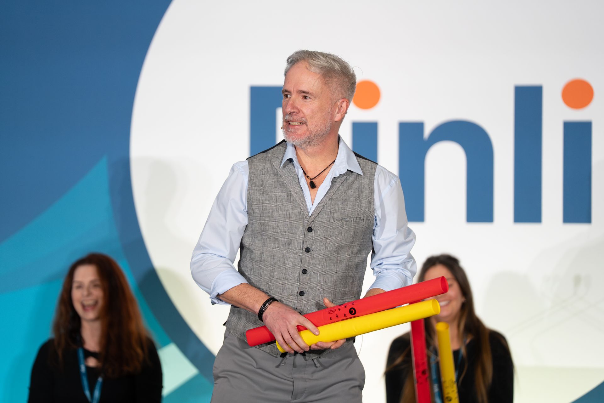 A speaker on stage holds colorful rhythm tubes, with two smiling audience members in the background.