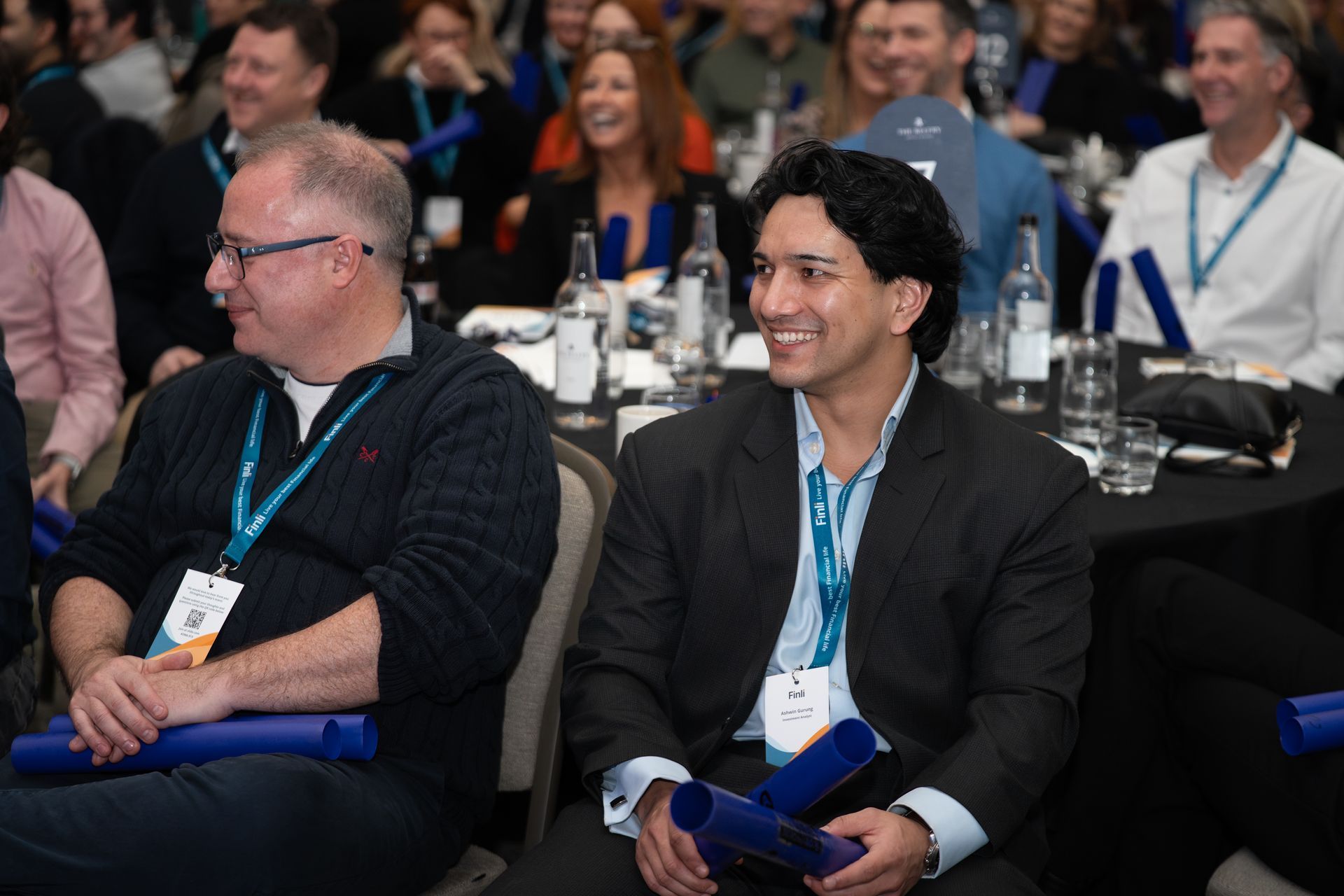 Two attendees smiling at a professional event, seated at tables with blue awards in hand.