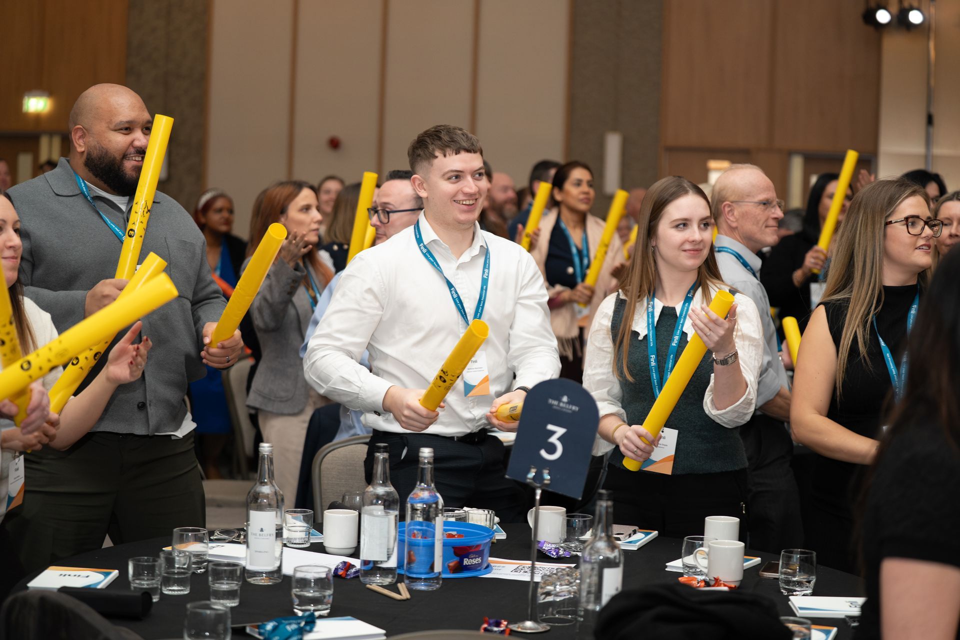 People at a conference table waving yellow foam sticks in a brightly lit ballroom.