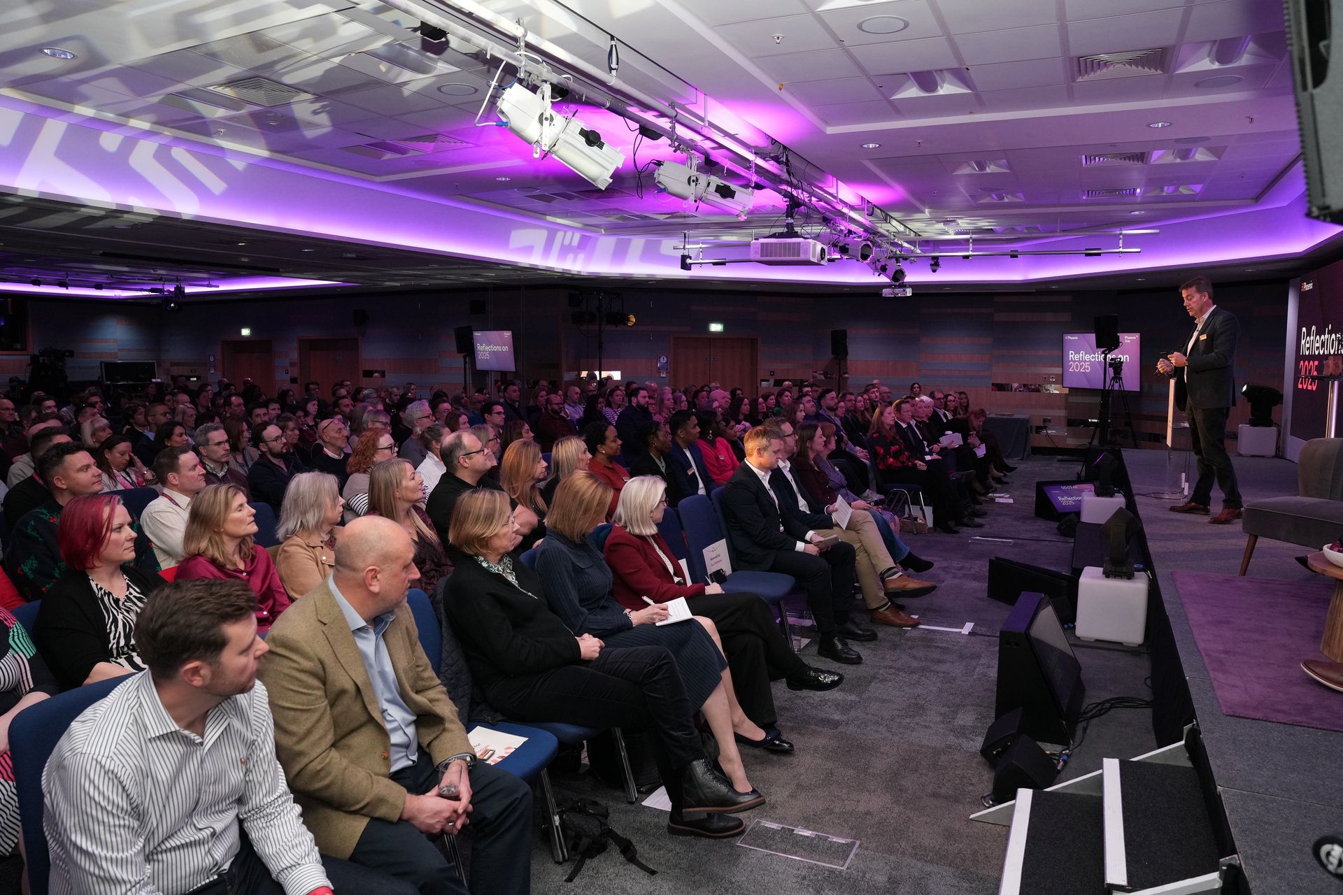 A speaker presents to a large audience seated in a dark, purple-lit conference room.