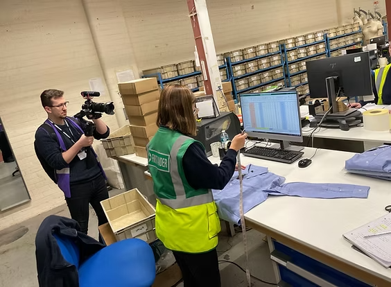 A person in a high-visibility vest measures a blue shirt on a desk while a videographer films them in a warehouse.