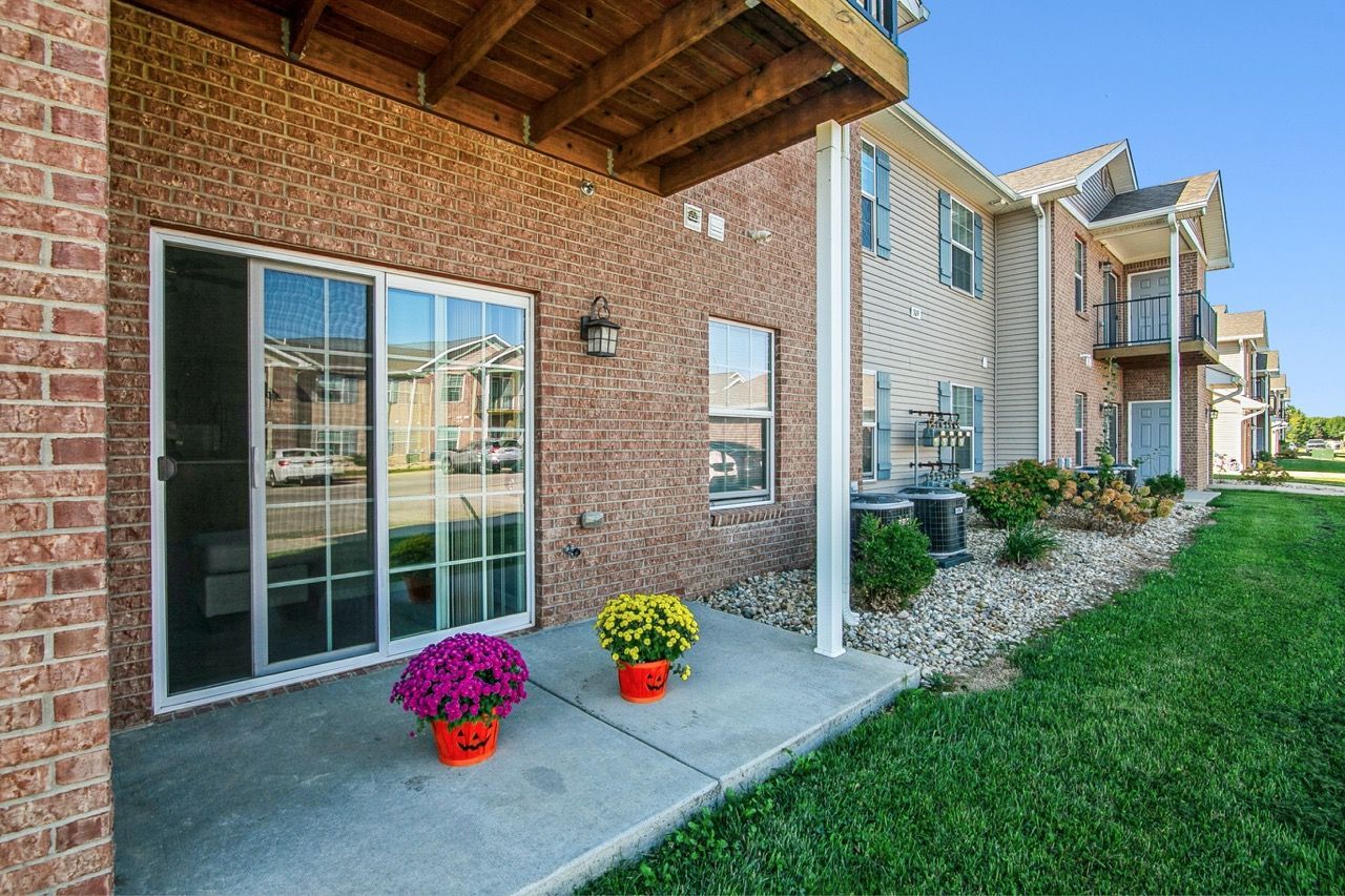 Brick apartment exterior with a sliding glass door, patio, and potted flowers. Green grass and blue sky.