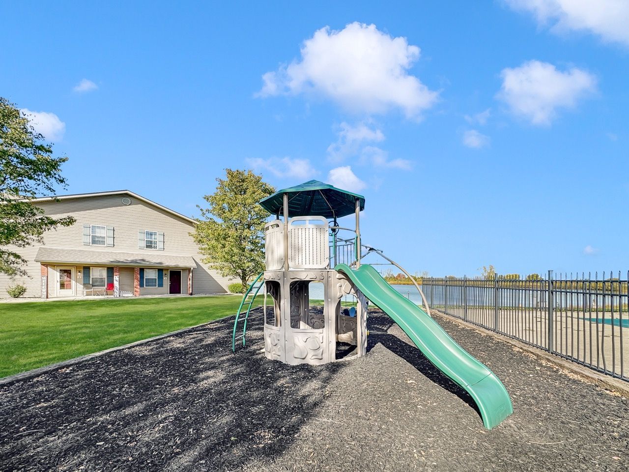 Playground with slide, adjacent to a building, fence, and green space, under a blue sky.