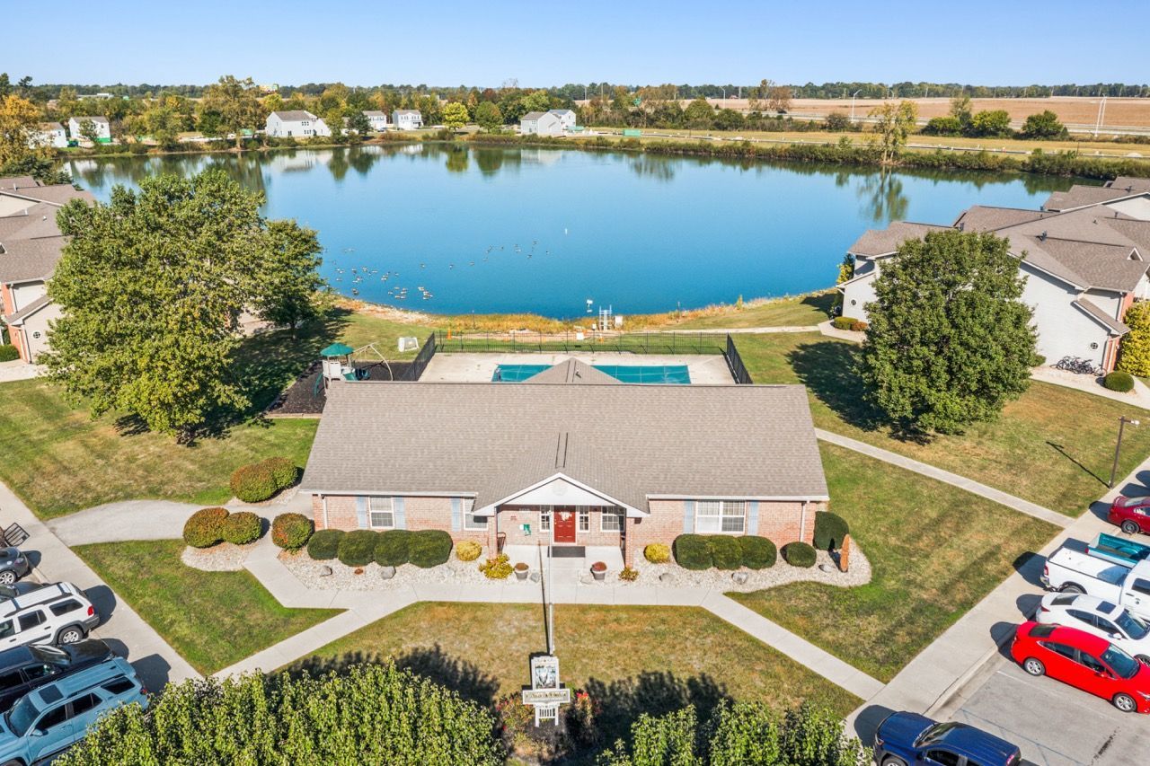 Aerial view of a building with pool and lake. Cars are parked. Green lawns and trees are around.