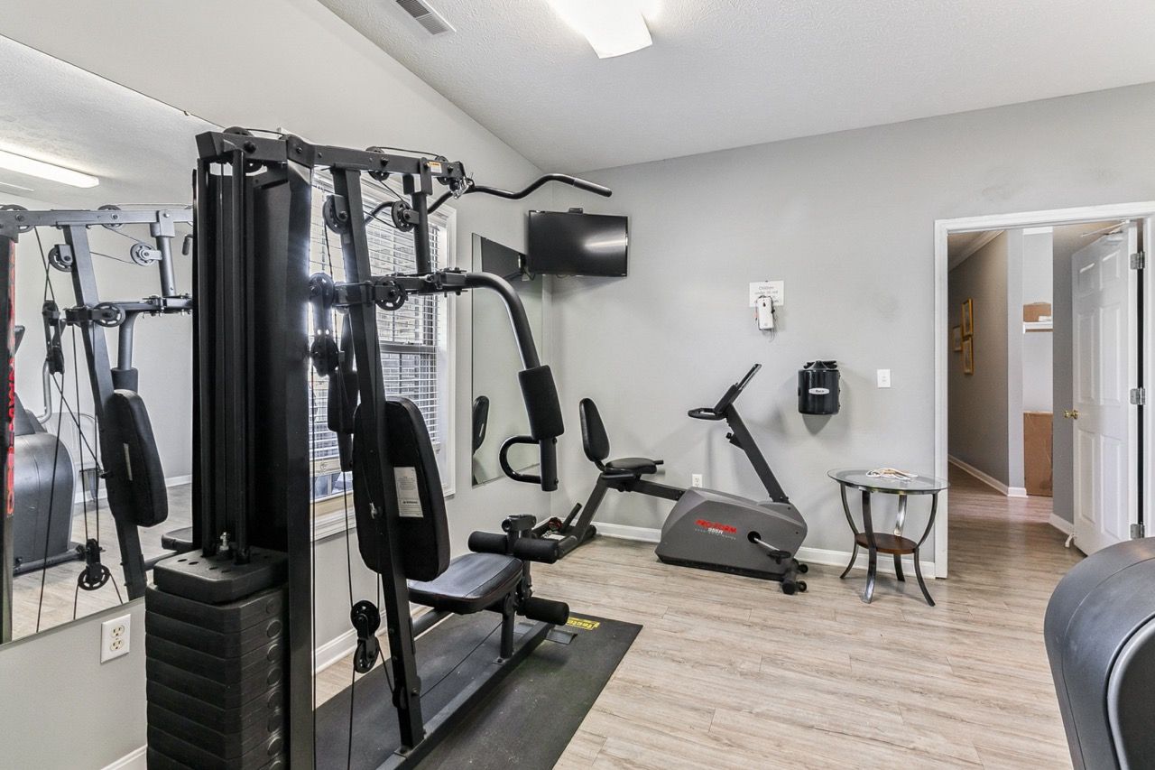 Gym with weight machine, stationary bike, and TV, gray walls, wood-look floor.