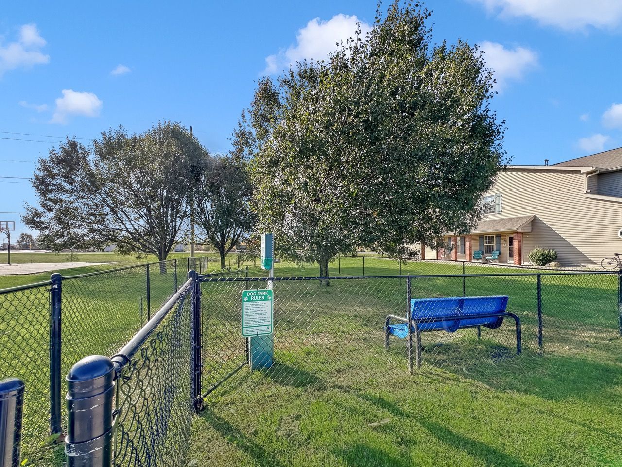 Fenced dog park with bench and sign under trees on a sunny day.