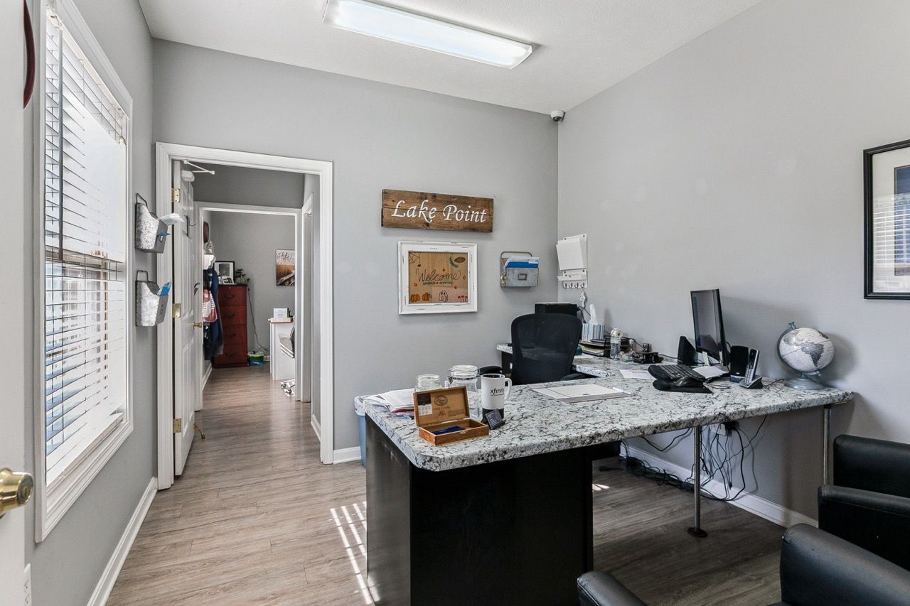Office interior with a desk, two chairs, and a hallway leading to other rooms. Gray walls, wood-look flooring.