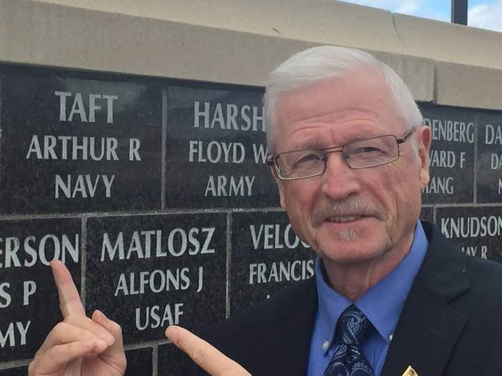 A man in a suit and tie points to a name on a wall