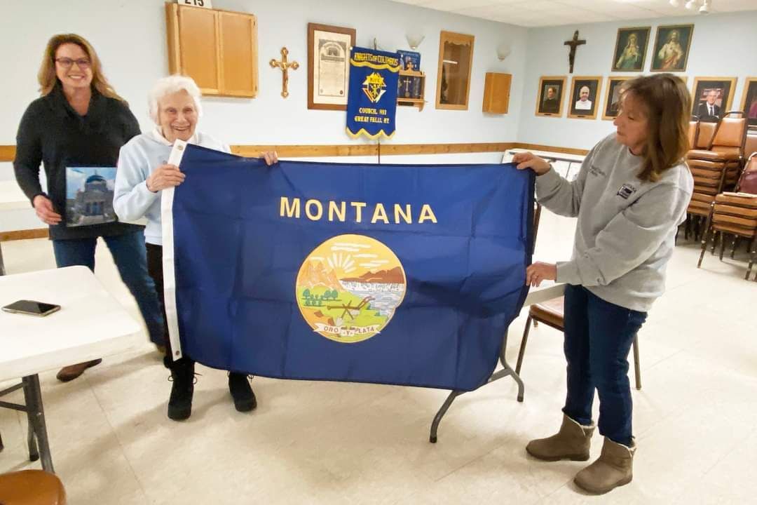 Three women are holding a montana flag in a room.