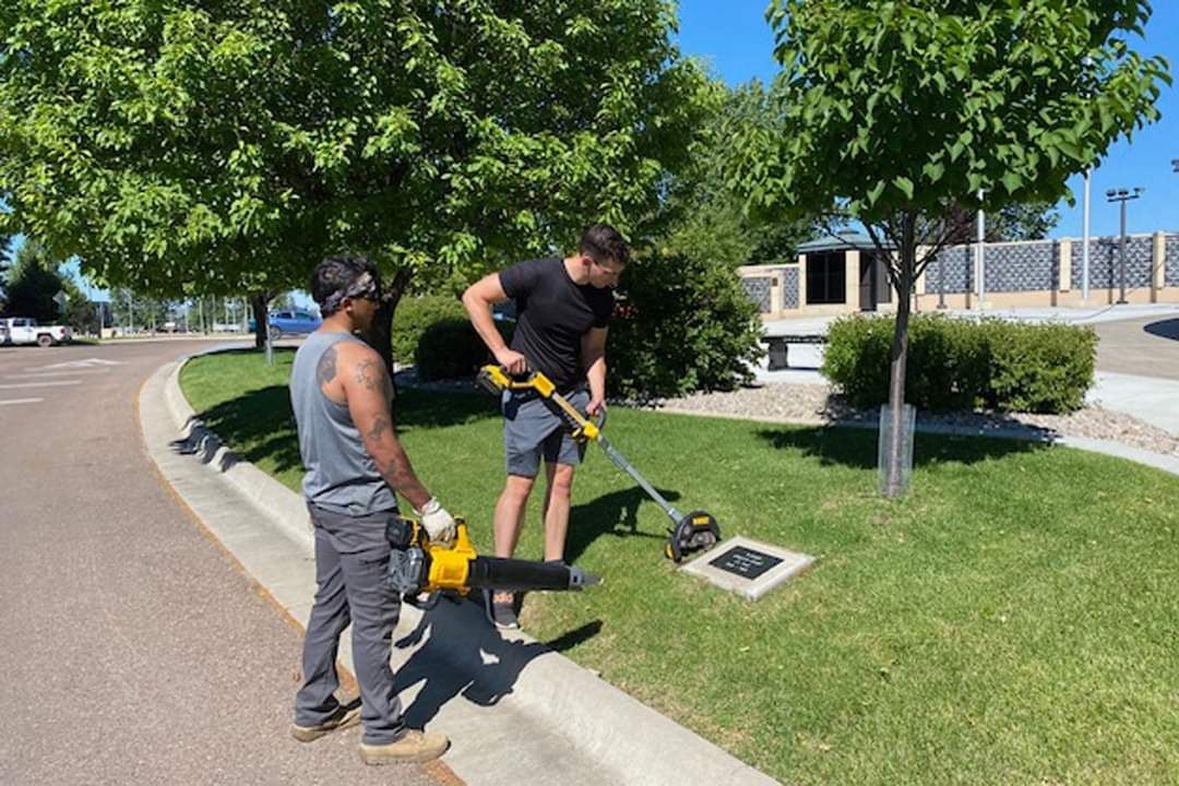 Two men are standing on the side of the road cutting grass with a lawn mower.