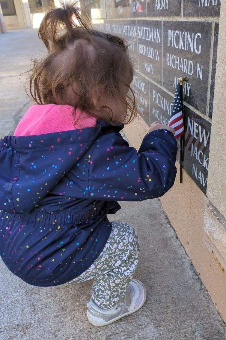 A little girl is kneeling down next to a wall holding an american flag.