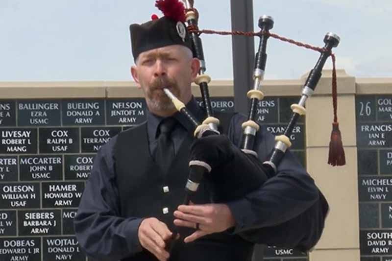 A man is playing a bagpipe in front of a wall with names on it