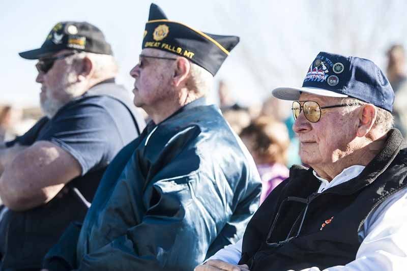 Three older men wearing hats and glasses are sitting in a crowd.