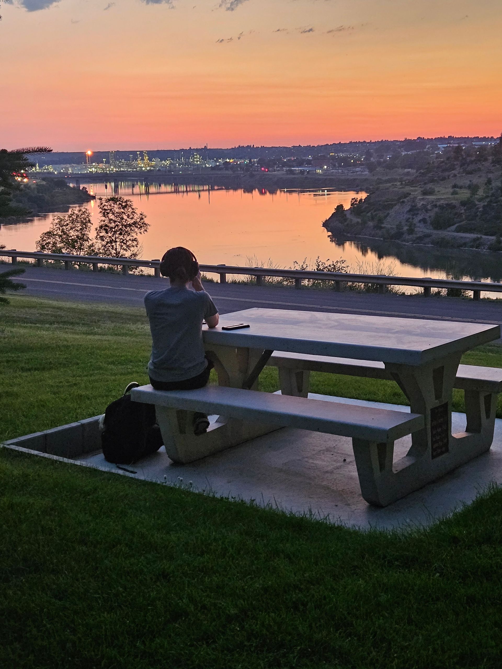 A man sits at a picnic table overlooking a lake at sunset