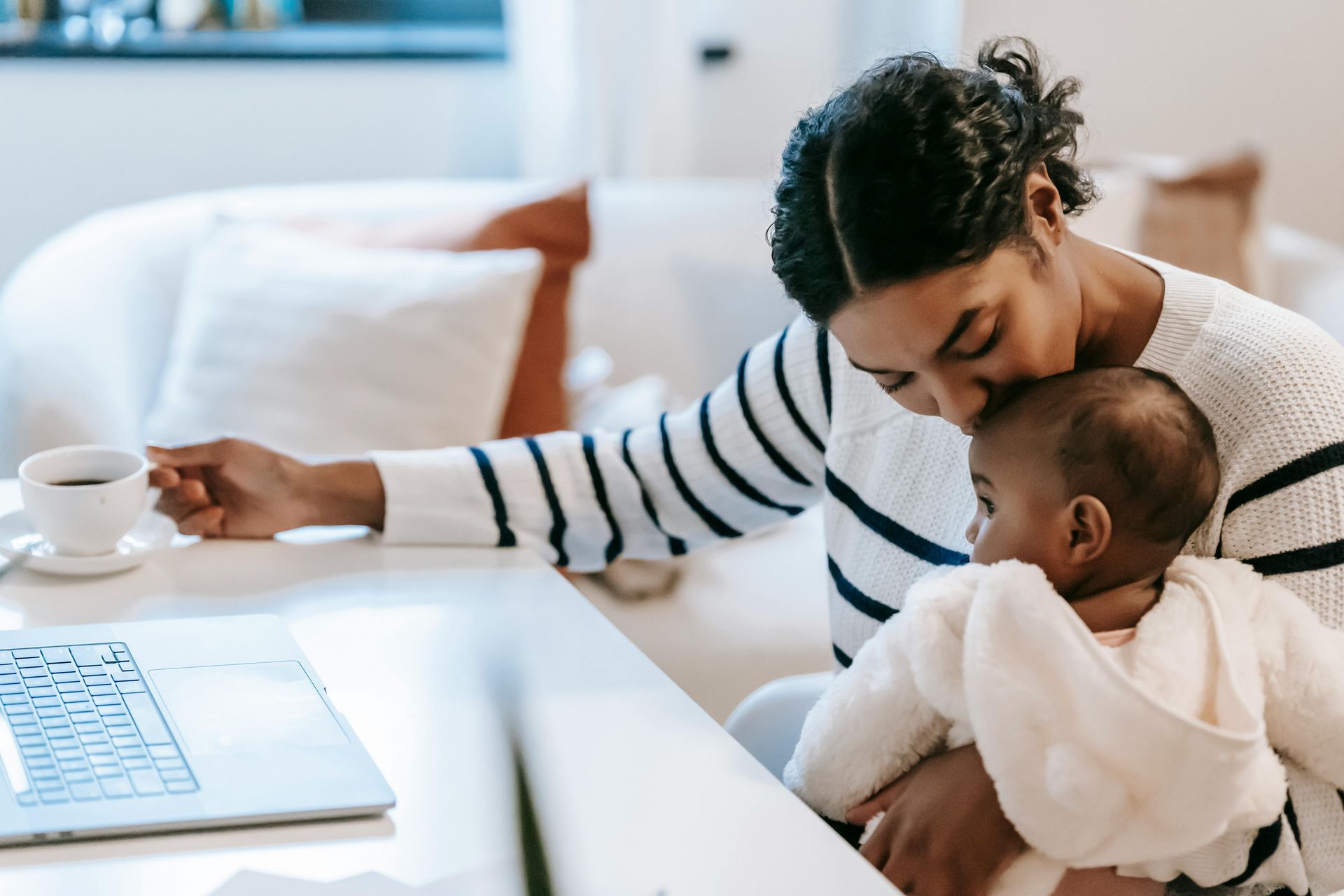 A woman is holding a baby while sitting at a desk with a laptop.