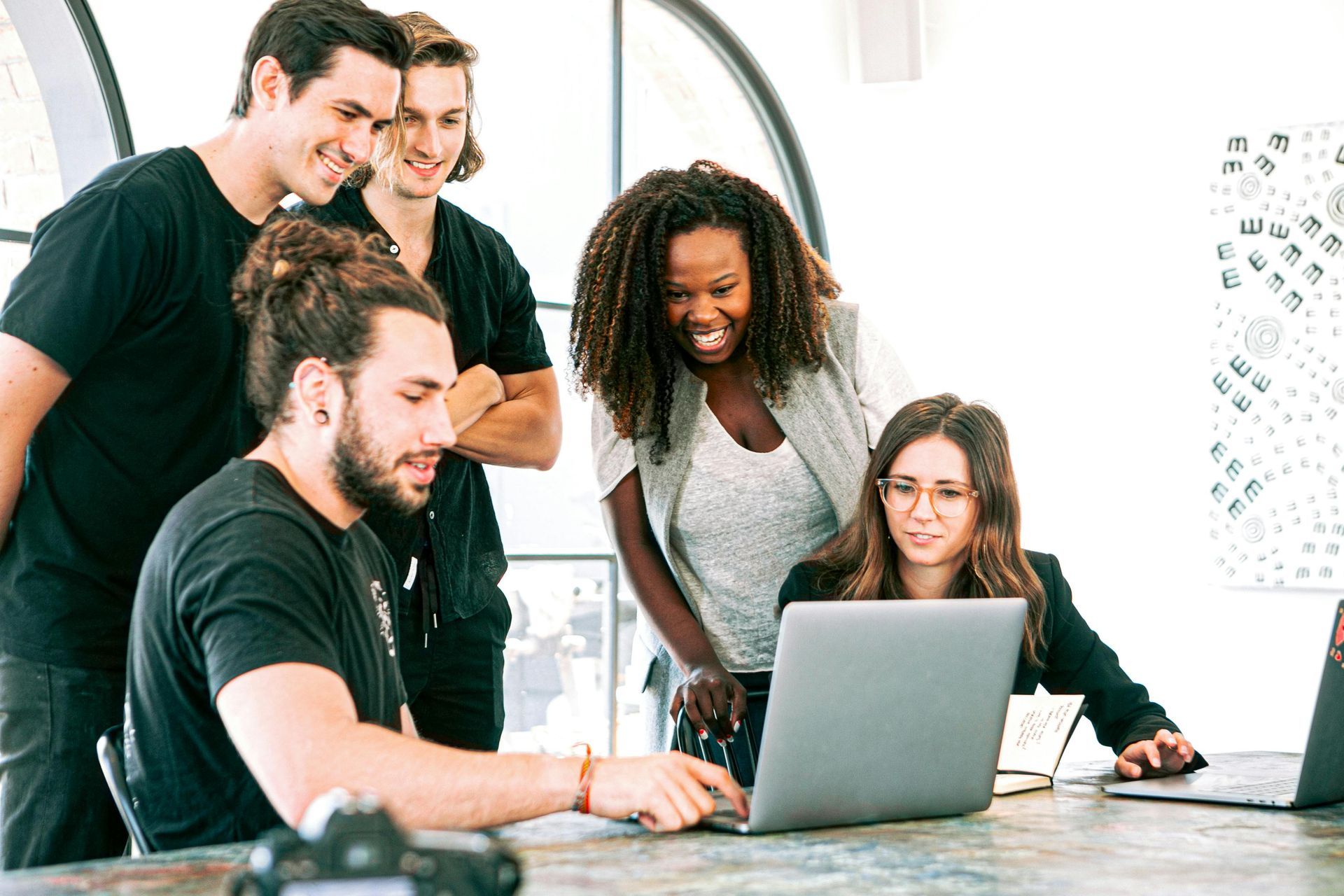 Group of people collaborating around a laptop, smiling. Bright office setting, light-skinned and Black individuals.
