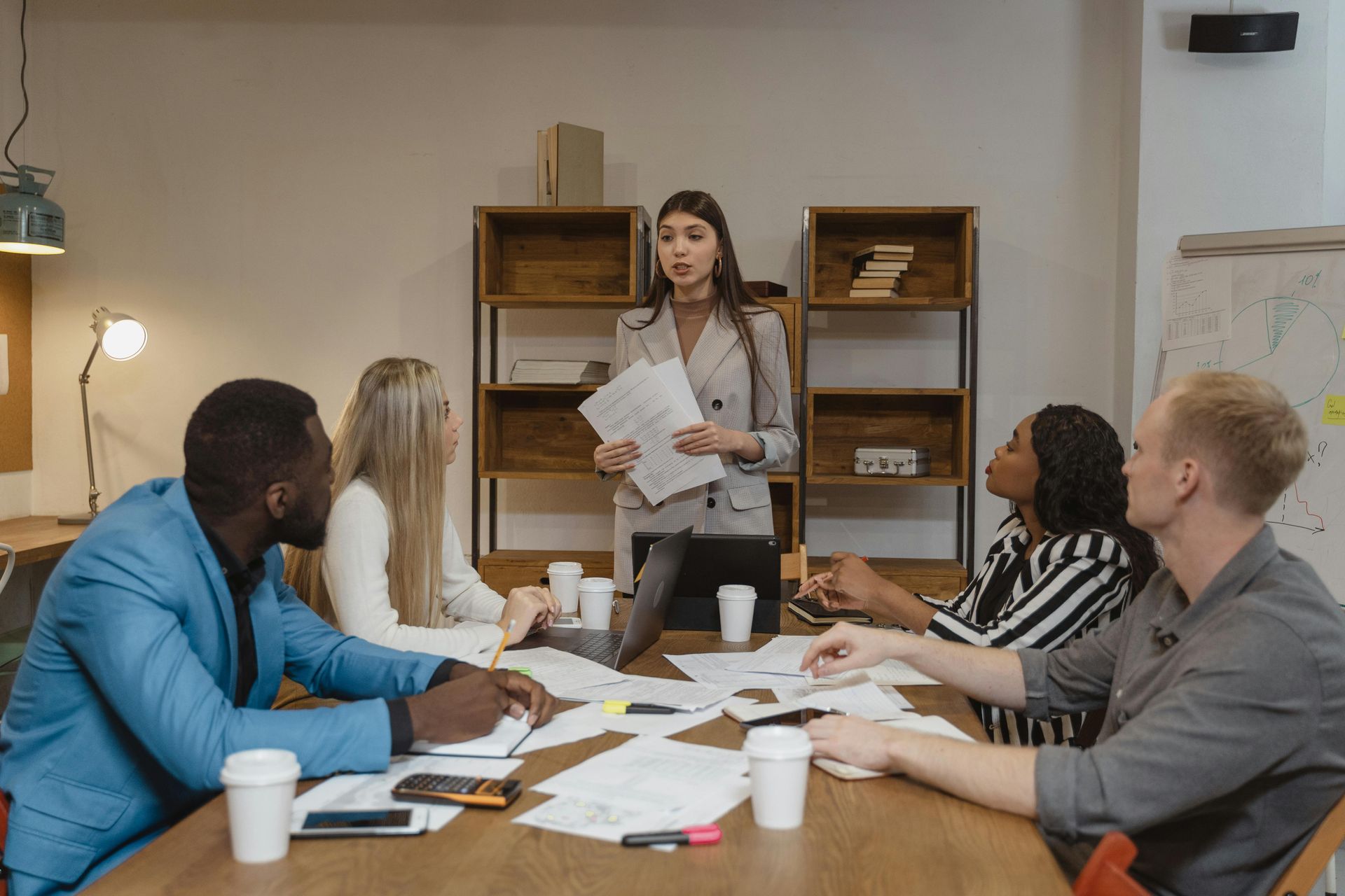 A woman in a blazer leads a meeting, holding papers.  Five colleagues sit around a table, looking on.