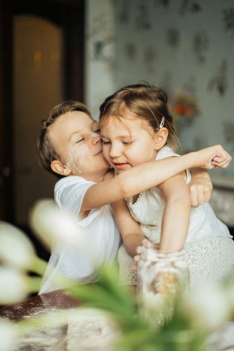 Two children hugging and smiling indoors, with soft light and blurred flowers in the foreground