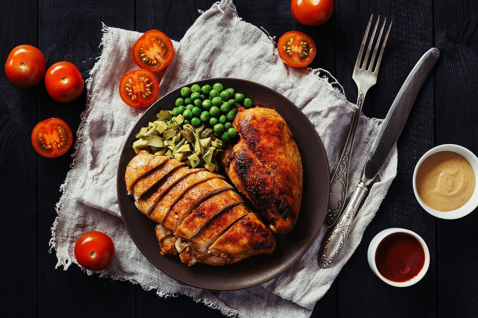 Grilled chicken breasts with peas and tomatoes on a plate, with dipping sauces and cutlery on a dark surface.