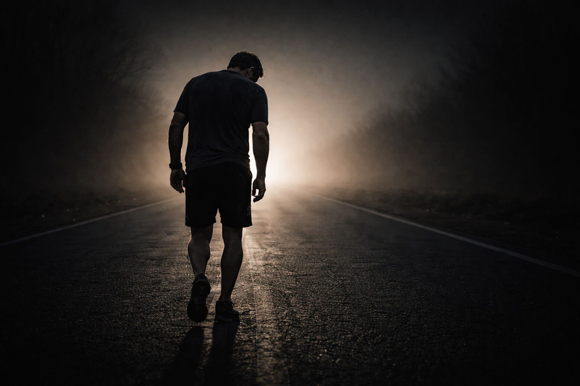 Man walks alone on a dark road towards a bright, foggy light.