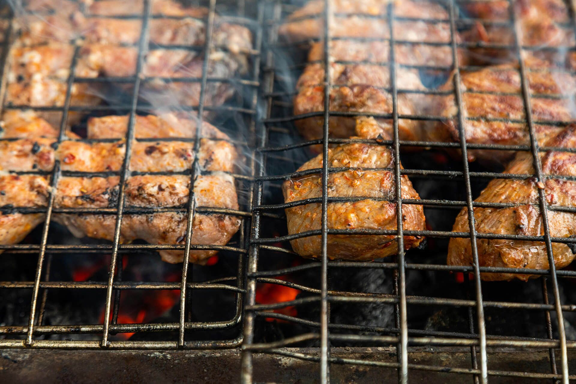 Chunks of seasoned meat cooking over hot coals in a metal wire grill basket.