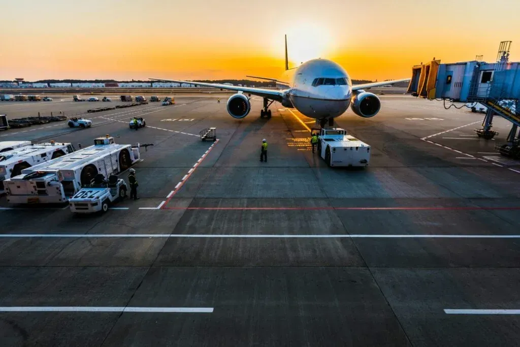 Airplane on tarmac at sunset, surrounded by support vehicles and a boarding bridge.