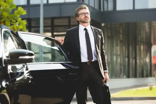 Man in suit, leaning on a black car, smiling. Building in background.