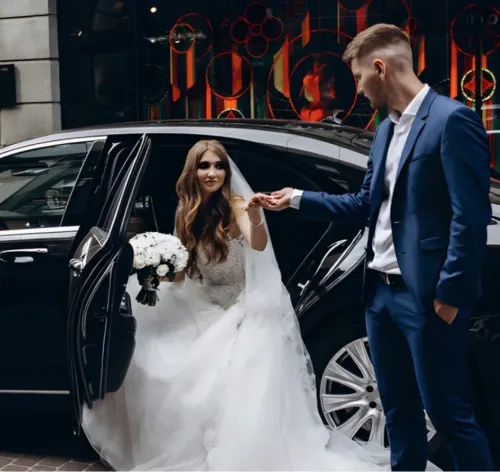 Bride exiting a black car, groom helping her. She wears a white gown, he wears a blue suit. Outside a building.