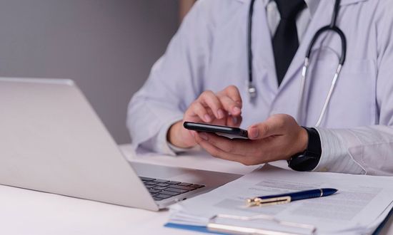 A healthcare professional in a white coat and stethoscope using a smartphone at a desk with a laptop and documents.