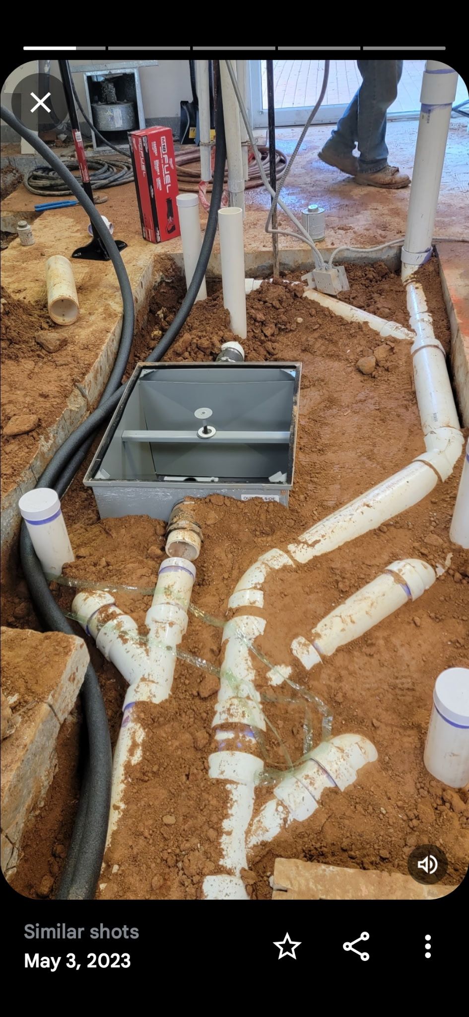 Plumber using a drill on pipes in a room with cinder block walls; wearing a tool belt.