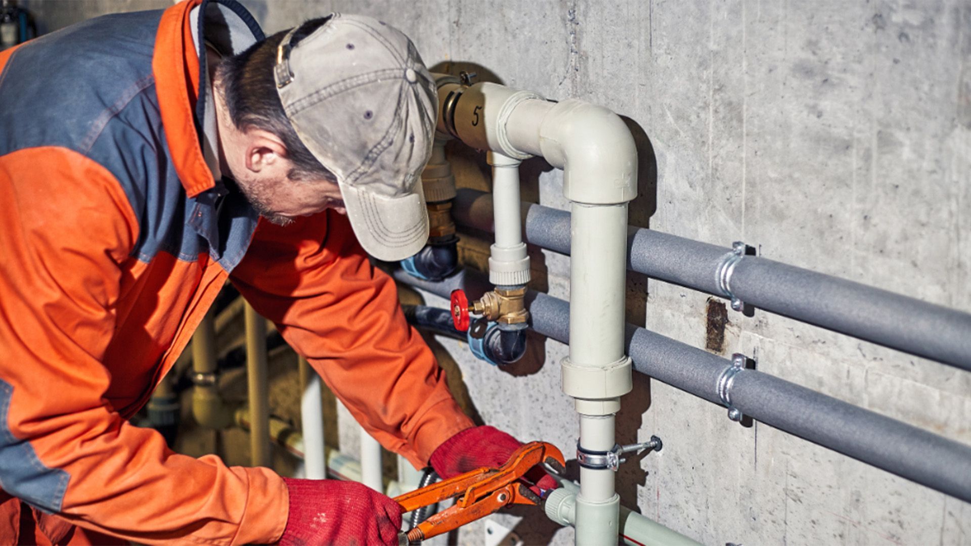 Plumber in orange jacket and red gloves working on pipes with pliers in a utility room.