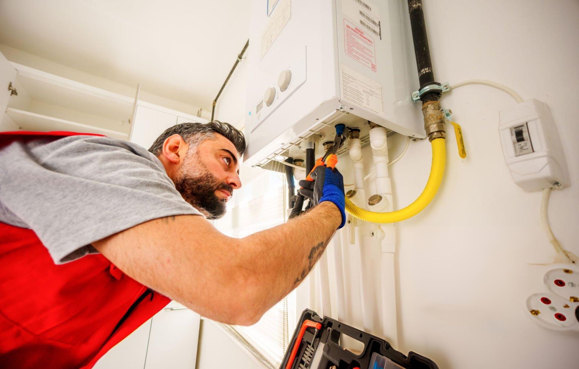 A man in a red vest repairs a water heater, holding a tool; white wall, yellow gas line.