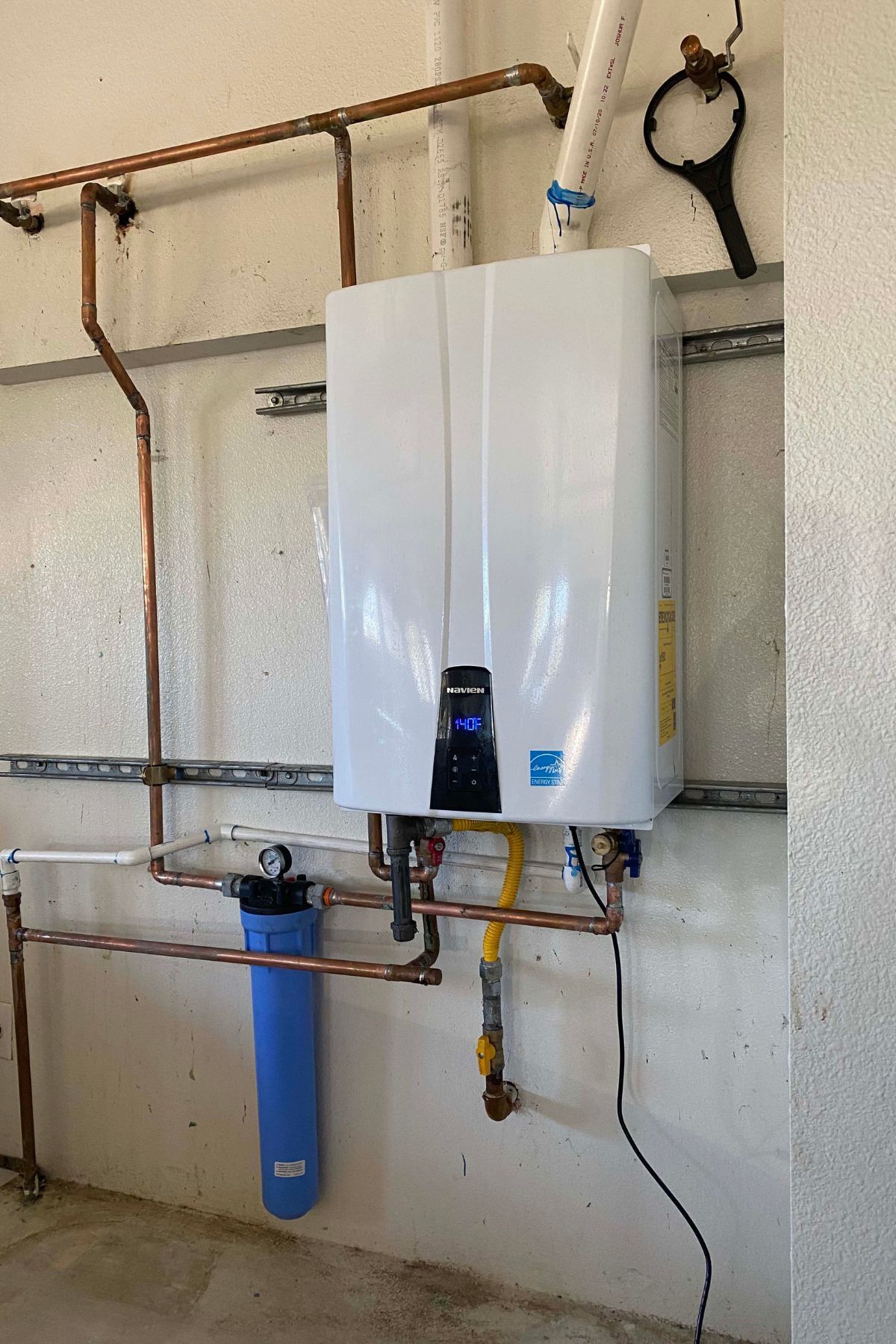 White tankless water heater with copper pipes and blue water filter on a concrete wall.