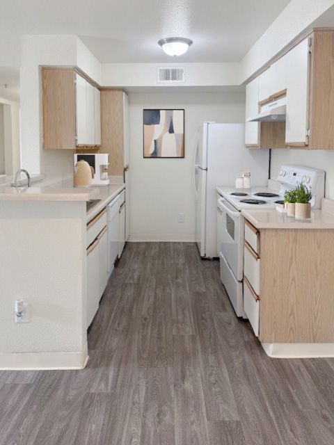 A galley kitchen with light wood cabinets, white appliances, and gray wood-look flooring under a central ceiling light.