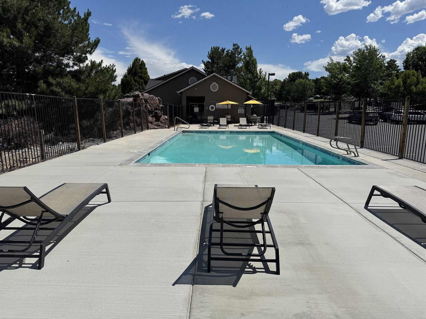 Pool area with lounge chairs, fenced pool, and a small building under a blue sky.