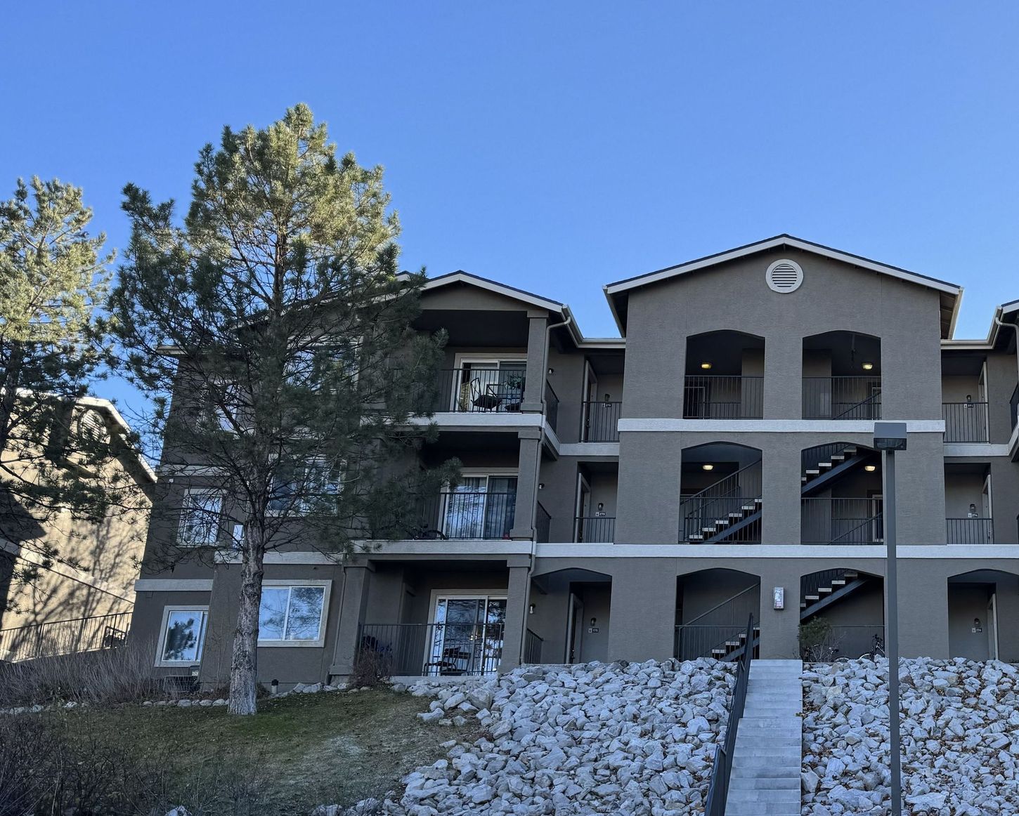Three-story apartment building with stucco exterior. Stone retaining wall in foreground; clear blue sky.