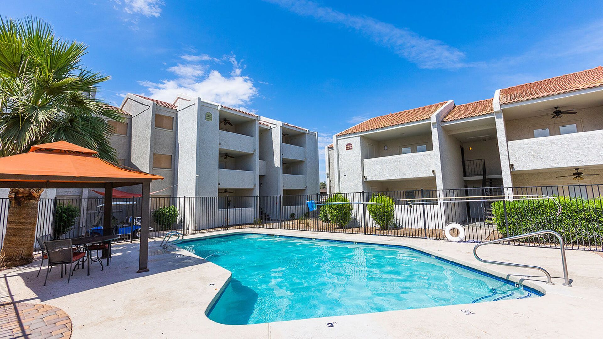 Apartment complex with a pool, gazebo, and palm trees under a blue sky.