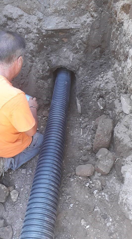 Uomo con camicia arancione che lavora con un tubo corrugato nero in una trincea di terra.