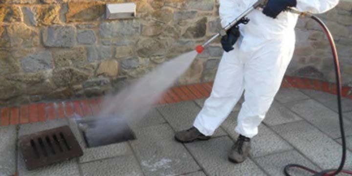 Persona in tuta protettiva bianca che utilizza un'idropulitrice per pulire uno scarico su un patio in pietra.