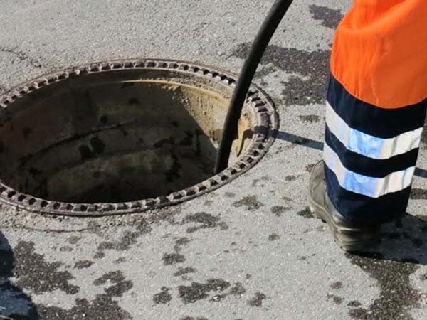 Un operaio con pantaloni arancioni e strisce riflettenti utilizza un attrezzo su un tombino in strada.