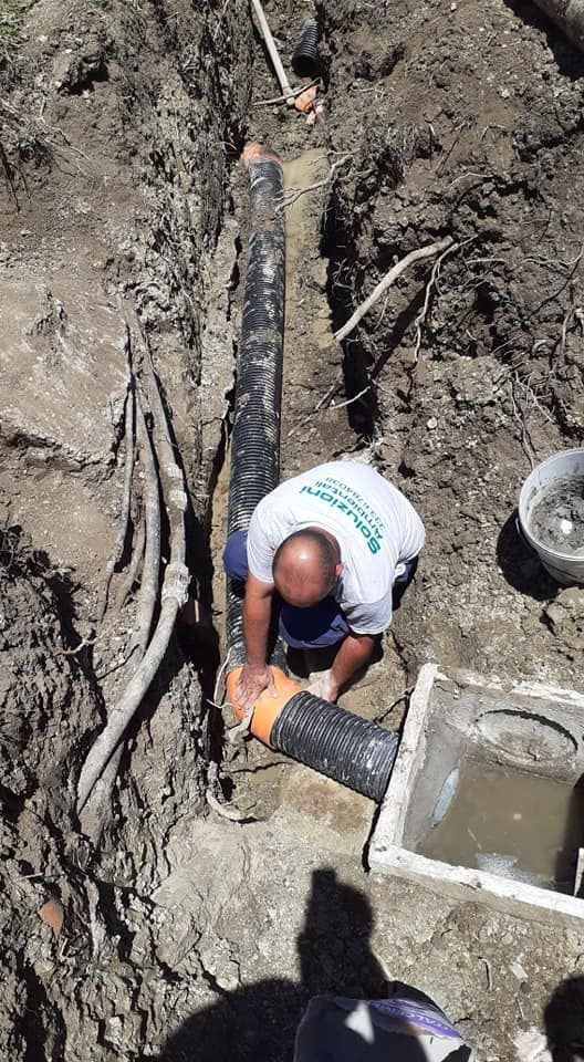 Uomo in una trincea che collega i tubi; sporcizia circostante, luce solare visibile.
