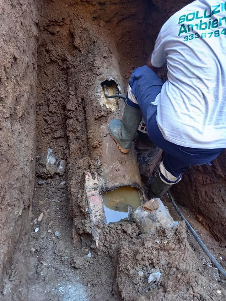 Una persona ispeziona una tubatura dell'acqua rotta in una trincea, con fango e acqua visibili.