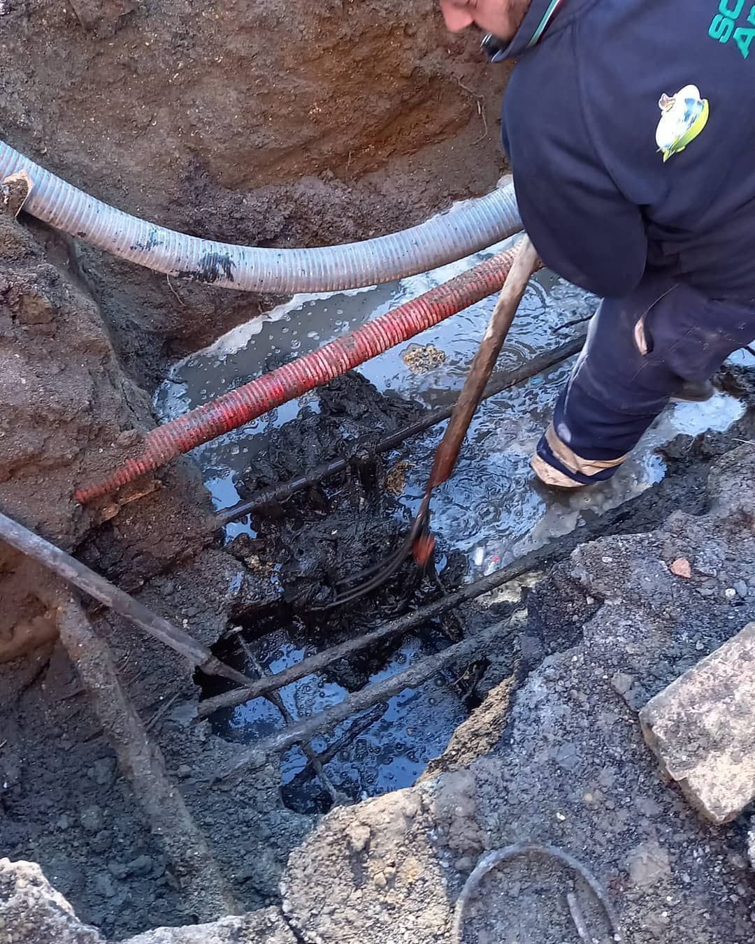 Un uomo in uniforme scura lavora in una trincea fangosa, esaminando cavi e acqua.