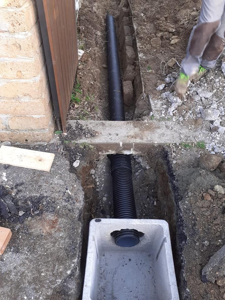 Tubo corrugato nero in trincea collegato alla scatola di drenaggio, accanto a un muro di mattoni e cemento.