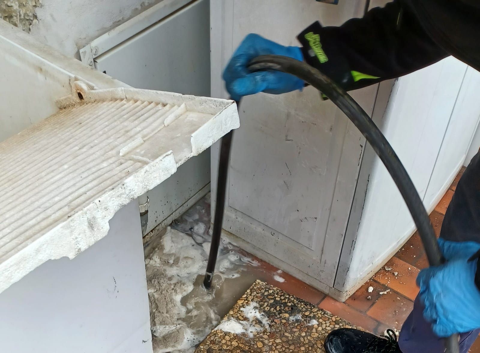 Persona che usa un tubo per pulire un'area allagata vicino a una sezione di muro rotta; sono visibili acqua e detriti.