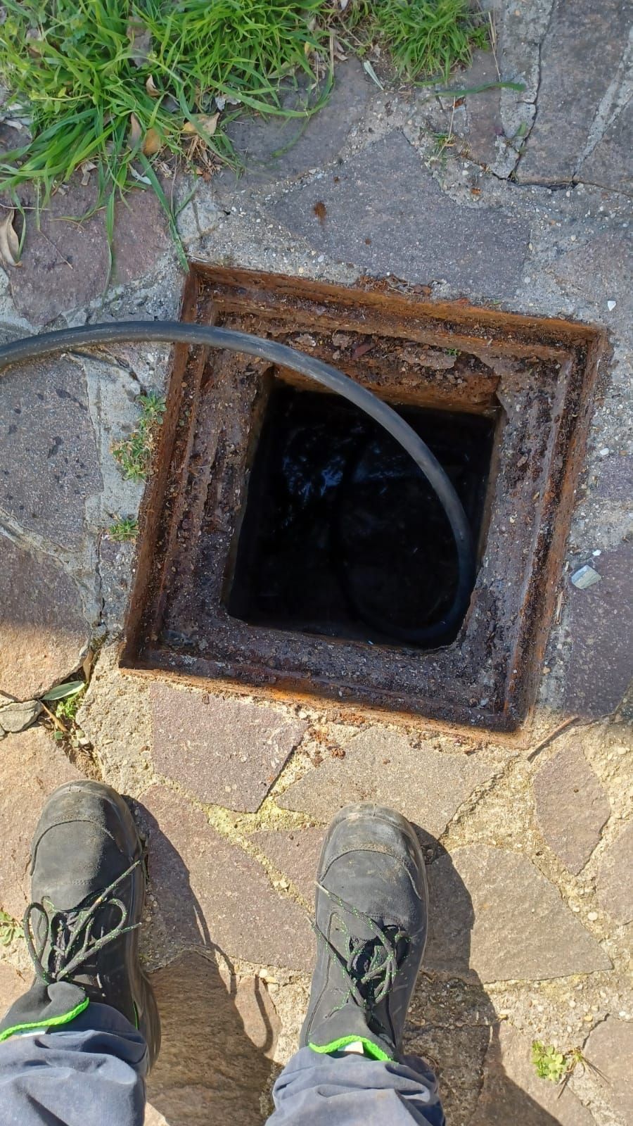 Persona in piedi vicino a una fogna a cielo aperto, con un cavo all'interno. Sono visibili il sentiero di ciottoli e l'erba verde.